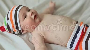 有趣的四个月大的婴儿躺在他的背上微笑。 穿着条纹短裤和帽子的婴儿